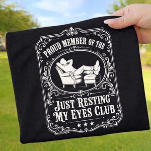 Könnte beinhalten: Schwarzes T-Shirt mit weißem Aufdruck. Der Aufdruck zeigt eine Person, die sich in einem Sessel zurücklehnt, mit dem Text "Proud Member of the Just Resting My Eyes Club". Das Design ist von Zierschmuck umrahmt.