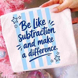 May include: A white t-shirt featuring a graphic design with vertical light blue stripes. The text in dark blue reads, "Be Like Subtraction and make a difference." The design is accented with several small, outlined star shapes in dark blue.