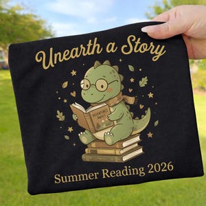 Könnte beinhalten: Schwarzes T-Shirt mit einem Cartoon-Dinosaurier, der ein Buch liest. Der Text auf dem Shirt lautet "Unearth a Story", "Adventures Await" und "Summer Reading 2026". Der Dinosaurier trägt eine Brille und einen Schal. Das Design ist in Gold und Grün gehalten.