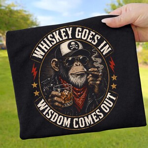 Puede incluir: Una camiseta negra con el gráfico de un chimpancé con gorra, gafas de sol y chaqueta de cuero. El chimpancé sostiene un vaso de whisky y un cigarro. El texto WHISKEY GOES IN WISDOM COMES OUT aparece en un diseño circular alrededor.