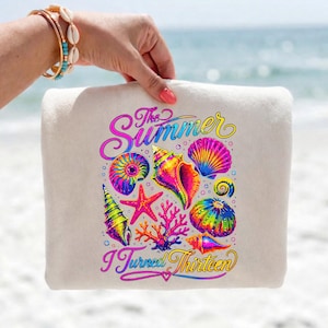 Könnte beinhalten: Ein beiges Strandtuch mit einem farbenfrohen Design aus Muscheln, einem Seestern und Korallen. Der Text "The Summer I Turned Thirteen" ist in einer mehrfarbigen Schrift dargestellt. Das Tuch wird vor einem verschwommenen Ozeanhintergrund gehalten.