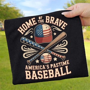 Könnte beinhalten: Schwarzes T-Shirt mit einem Baseball-Motiv. Das Design zeigt eine Baseballkappe, gekreuzte Schläger und einen Baseball, alles in patriotischen Rot-, Weiß- und Blautönen. Der Text lautet "HOME OF THE BRAVE" und "AMERICA'S PASTIME BASEBALL."