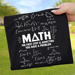 May include: A black t-shirt featuring white printed mathematical formulas and graphs. The centre displays the word MATH with a triangle replacing the letter A, followed by the text THE FIRST STEP IS ADMITTING YOU HAVE A PROBLEM.