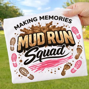 Puede incluir: Camiseta blanca con las palabras "MAKING MEMORIES MUD RUN Squad" en letras negras y doradas. El diseño incluye salpicaduras de barro marrón, huellas rosas y marrones y una raya rosa. La camiseta se sostiene sobre un fondo verde.