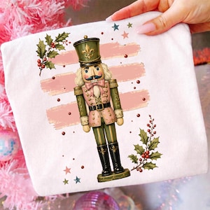 Könnte beinhalten: Ein weißes T-Shirt mit einer Grafik eines Nussknacker-Soldaten in einer rosa und olivgrünen Uniform. Das Design enthält Stechpalmenblätter mit roten Beeren, kleine verstreute rote Punkte und mehrere kleine Sterne in Rosa, Blau und Grün.