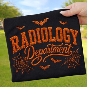 Könnte beinhalten: Ein schwarzes T-Shirt mit orangefarbener Stickerei, die den Text RADIOLOGY Department in einer Mischung aus Block- und Schreibschrift zeigt. Das Design umfasst gestickte orangefarbene Fledermäuse und Spinnennetze um den Text herum.