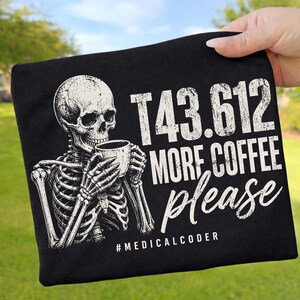 Könnte beinhalten: Ein schwarzes T-Shirt mit einer weißen Grafik eines Skeletts, das eine Kaffeetasse hält. Der Text lautet T43.612 MORE COFFEE please und #MEDICALCODER. Das Design hat ein abgenutztes, strukturiertes Aussehen auf der Vorderseite.