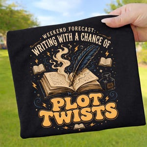 Könnte beinhalten: Ein schwarzes T-Shirt mit einer Grafik eines aufgeschlagenen Buches, auf dem eine Feder liegt, umgeben von Rauch, kleinen Sternen und Blitzen. Der Text lautet: WEEKEND FORECAST: WRITING WITH A CHANCE OF PLOT TWISTS und INK.