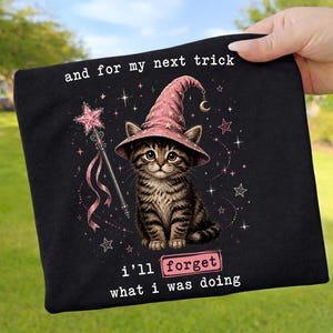 Könnte beinhalten: Schwarzes T-Shirt mit einer getigerten Katze, die einen rosa Zaubererhut trägt und einen Sternenstab hält. Der Text lautet "and for my next trick, I'll forget what I was doing". Sterne und ein Mond sind ebenfalls im Design enthalten.
