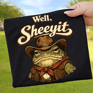 Könnte beinhalten: Schwarzes T-Shirt mit einer Cartoon-Frosch, der einen Cowboyhut, eine Weste und ein rotes Halstuch trägt. Der Text "Well, Sheeeyit" steht über dem Frosch. Der Frosch hat einen ernsten Gesichtsausdruck.