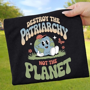 Könnte beinhalten: Schwarzes T-Shirt mit dem Text "DESTROY THE PATRIARCHY NOT THE PLANET". Das Design zeigt einen Cartoon-Erdcharakter mit roter Kappe, der auf Gras mit Blumen und einem Schmetterling sitzt.