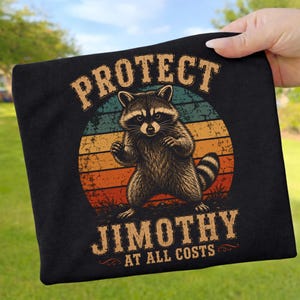 Könnte beinhalten: Ein schwarzes T-Shirt mit einer Grafik eines Waschbären vor einem verwaschenen, mehrfarbigen Sonnenuntergangskreis. Der Text "PROTECT JIMOTHY AT ALL COSTS" ist in einer fetten Vintage-Schrift über und unter der Waschbären-Illustration gedruckt.