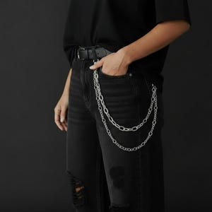 May include: A silver chain belt with two strands hangs from the belt loop of black ripped jeans. The jeans are paired with a black belt and a black top. The image is set against a dark background.