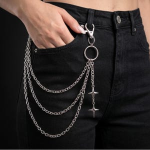 May include: A silver-toned metal wallet chain attached to a belt loop with a lobster clasp. The accessory features three draped chain layers connected to a circular ring, which also holds two dangling four-pointed star charms on black jeans.