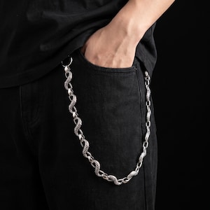 Könnte beinhalten: Eine silberfarbene Metall-Geldbörsenkette mit einer Reihe von strukturierten, schlangenförmigen Gliedern. Die Kette ist an der Tasche einer schwarzen Jeans befestigt und zeigt ein detailliertes, schuppiges Oberflächenmuster auf jedem einzelnen Glied.