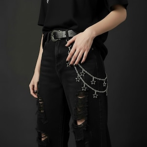 May include: Black distressed jeans with a silver chain belt featuring star charms. The belt is paired with a black leather belt with a decorative buckle. The jeans have ripped details.