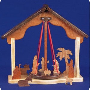 May include: Wooden nativity scene with a rotating base and red ribbons. The figures include Mary, Joseph, baby Jesus, a donkey, a sheep, a shepherd, and an angel. The scene is set in a wooden stable with a palm tree.