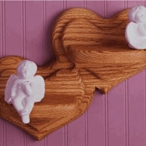 May include: Two heart-shaped wooden shelves with a natural finish are mounted on a pink wall. Each shelf has a small, white cherub figurine. The shelves are designed to hold small decorative items.