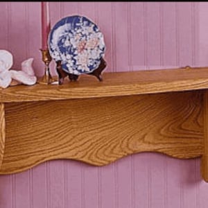 Puede incluir: Un estante de madera con un borde frontal curvo y soportes decorativos. El estante está hecho de madera marrón claro y tiene un acabado natural. Está montado en una pared rosa y tiene un pequeño plato decorativo y una figurita en la parte superior.