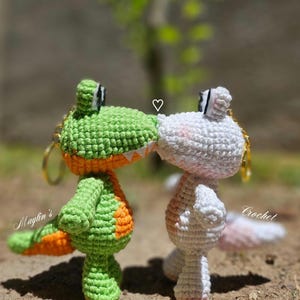Puede incluir: Dos llaveros de ganchillo con forma de cocodrilo, uno verde con vientre naranja y otro blanco con vientre rosa, enfrentados con un peque&ntilde;o s&iacute;mbolo de coraz&oacute;n blanco entre ellos. El texto Maylin's Crochet es visible en la imagen.