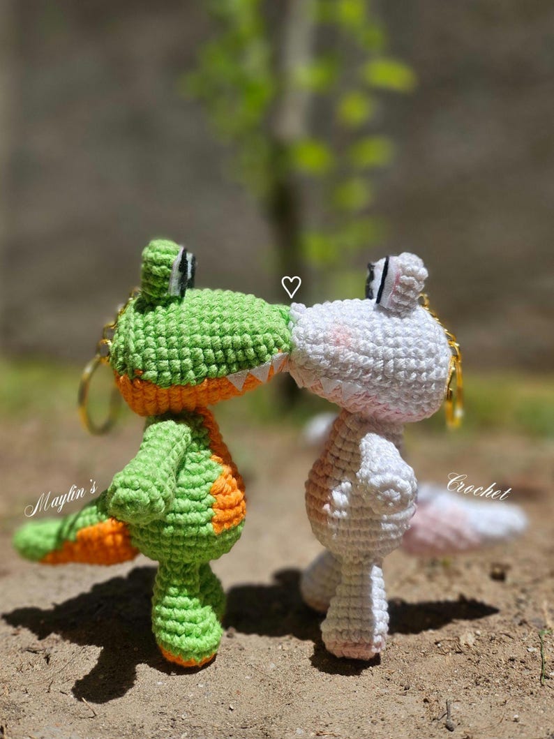 Puede incluir: Dos llaveros de ganchillo con forma de cocodrilo, uno verde con vientre naranja y otro blanco con vientre rosa, enfrentados con un peque&ntilde;o s&iacute;mbolo de coraz&oacute;n blanco entre ellos. El texto Maylin's Crochet es visible en la imagen.