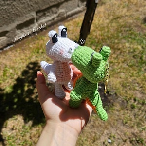 Puede incluir: Dos peque&ntilde;as figuras de dinosaurios de ganchillo hechas a mano, una blanca y otra verde, frente a frente como si se besaran. El dinosaurio verde tiene un llavero de metal dorado en la espalda. El texto Maylin's Crochet es visible.