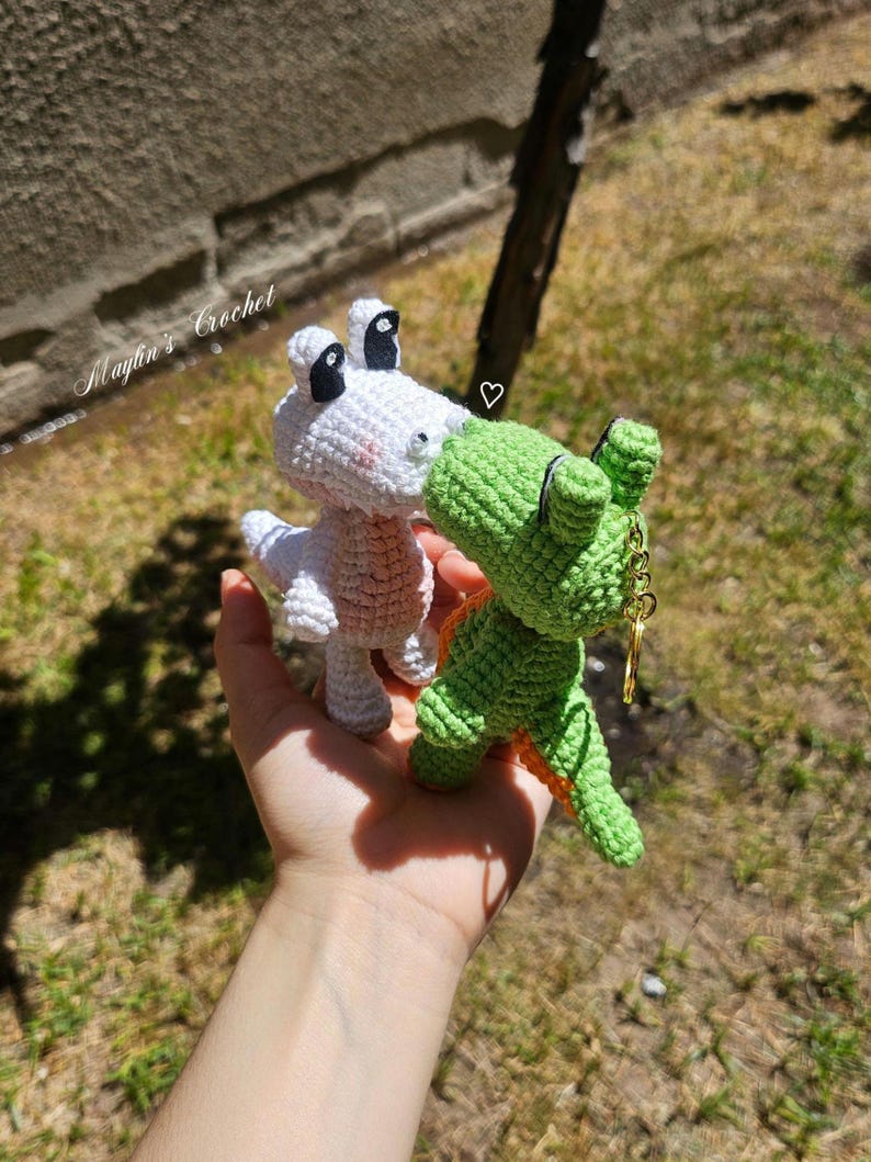 Puede incluir: Dos peque&ntilde;as figuras de dinosaurios de ganchillo hechas a mano, una blanca y otra verde, frente a frente como si se besaran. El dinosaurio verde tiene un llavero de metal dorado en la espalda. El texto Maylin's Crochet es visible.