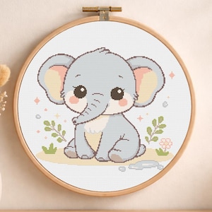 Pode incluir: Bordado em ponto cruz de um elefante bebé num bastidor de madeira. O elefante é cinzento com detalhes cor-de-rosa nas orelhas e bochechas. O desenho inclui pequenas flores e folhas num fundo amarelo claro.