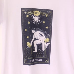Peut inclure: T-shirt blanc avec un motif représentant la carte de tarot L'Étoile. La carte représente une femme versant de l'eau de deux jarres, avec une étoile au-dessus de sa tête. La carte est encadrée d'or et porte le chiffre romain XVII et les mots "L'Étoile" imprimés dessus.
