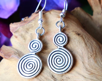 May include: Silver earrings with a double spiral design. The earrings are hanging from a wooden surface with purple flowers in the background.