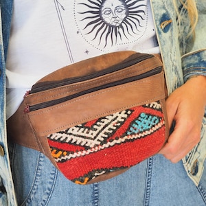 Può includere: Una marsupio in pelle marrone con una patta a motivi rossi, blu e bianchi. Il marsupio è indossato sopra un paio di jeans blu e una maglietta bianca con un'illustrazione in bianco e nero del sole e della luna con il testo "The Tower".