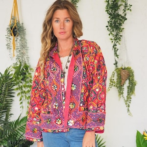 May include: A brightly coloured, embroidered jacket with a floral pattern in red, purple, and pink. The jacket has long sleeves and a collared neckline. The model is wearing light blue jeans.