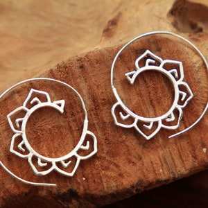 May include: A pair of silver-colored spiral earrings with a floral design. The earrings are set against a natural wood background, highlighting their intricate details and bohemian style. These earrings are a unique accessory.
