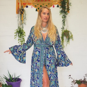 May include: A long, flowing blue dress with a paisley pattern. The dress features wide bell sleeves and a plunging neckline. The fabric has teal and gold accents. The dress is floor-length and has a high slit in the front.