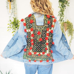 May include: Light blue denim jacket with a colorful embroidered back panel. The panel features a geometric design in black, green, and red, accented with red pom-poms. The jacket is paired with light blue jeans, creating a casual, bohemian look.