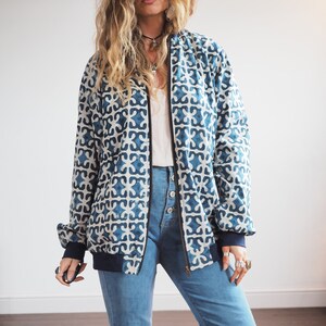 May include: A long-sleeve bomber jacket featuring a repeating blue and white geometric pattern, a black front zipper, and solid dark blue ribbed cuffs and hem, paired with high-waisted blue denim jeans with a visible button-front closure.
