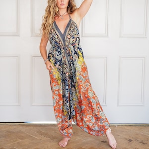 Op de afbeelding: Een vrouw draagt een kleurrijke, geprinte jumpsuit met een halternek. De jumpsuit heeft een paisley-ontwerp in tinten blauw, oranje en geel. De vrouw staat voor een witte deur met houten lambrisering.