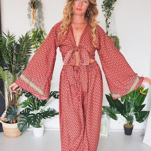 May include: A red and blue patterned two-piece outfit with a long-sleeved top and wide-leg trousers. The top has a tie at the waist and the trousers have a drawstring.