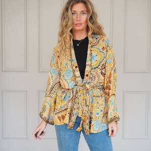 May include: A patterned kimono in shades of yellow, blue, and brown. The kimono has a tie waist and long sleeves. The garment is worn over a black top and paired with blue jeans. The background is a white door.