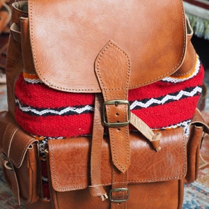 May include: A brown leather backpack with a red, white and black woven pattern on the front. The backpack has two front pockets and a buckle closure.