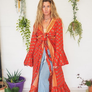 May include: A long red floral print maxi dress with a tie waist and bell sleeves. The dress has a ruffled hem and is worn with blue jeans.