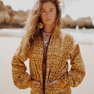 Puede incluir: Chaqueta bomber de color mostaza con un patrón repetido de formas abstractas. La chaqueta tiene una cremallera negra y bolsillos. La modelo está de pie en una playa con el océano al fondo. Los puños y el dobladillo de la chaqueta son de color mostaza liso.