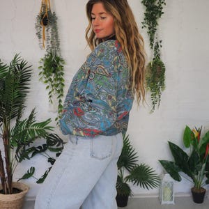 Op de afbeelding: Een paisley-gepatroneerde jas in blauw, groen, rood en geel, gecombineerd met lichtblauwe jeans. De jas heeft een zwarte kraag. Het model staat voor een witte muur met hangplanten en potgroen.