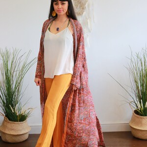 Puede incluir: Una mujer lleva un kimono largo y fluido con estampado paisley, un top blanco de tirantes y pantalones amarillos mostaza. Est&aacute; de pie frente a una pared blanca con un atrapasue&ntilde;os colgado.