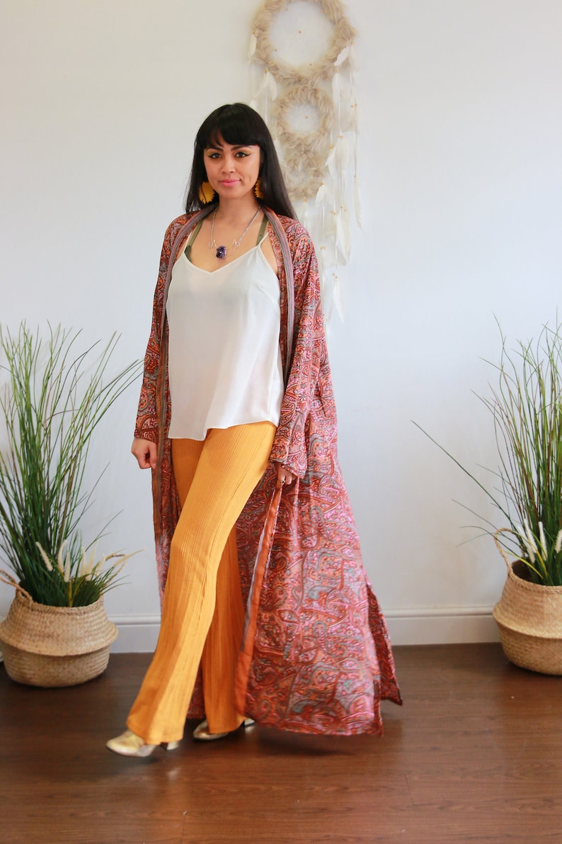 Puede incluir: Una mujer lleva un kimono largo y fluido con estampado paisley, un top blanco de tirantes y pantalones amarillos mostaza. Est&aacute; de pie frente a una pared blanca con un atrapasue&ntilde;os colgado.
