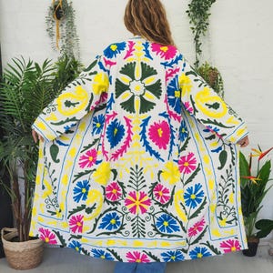 May include: A white embroidered coat with a long sleeve and a vibrant floral pattern in yellow, pink, blue, and green. The coat features a detailed design with a symmetrical arrangement of flowers and leaves, creating a colorful and eye-catching garment.