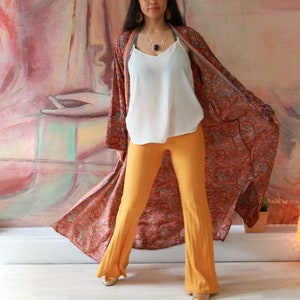 Puede incluir: Una mujer con un kimono largo, fluido, con estampado de paisley, un top blanco de tirantes y pantalones anchos de color mostaza. Est&aacute; de pie frente a una gran pintura.