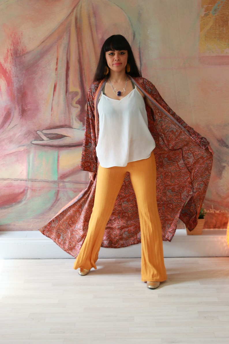 Puede incluir: Una mujer con un kimono largo, fluido, con estampado de paisley, un top blanco de tirantes y pantalones anchos de color mostaza. Est&aacute; de pie frente a una gran pintura.