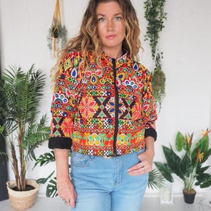 May include: A colorful, patterned jacket with a black zipper and cuffs. The jacket features a vibrant mix of red, yellow, blue, and green floral and geometric designs. The person is wearing light blue jeans.