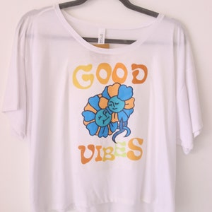 Può includere: Maglietta bianca corta con una grafica di due fiori blu con facce e il testo "GOOD VIBES" in arancione e giallo.